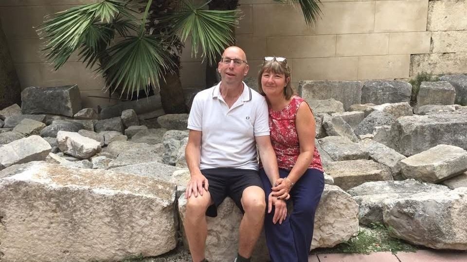 A couple sit on a large stone with a palm tree behind them.